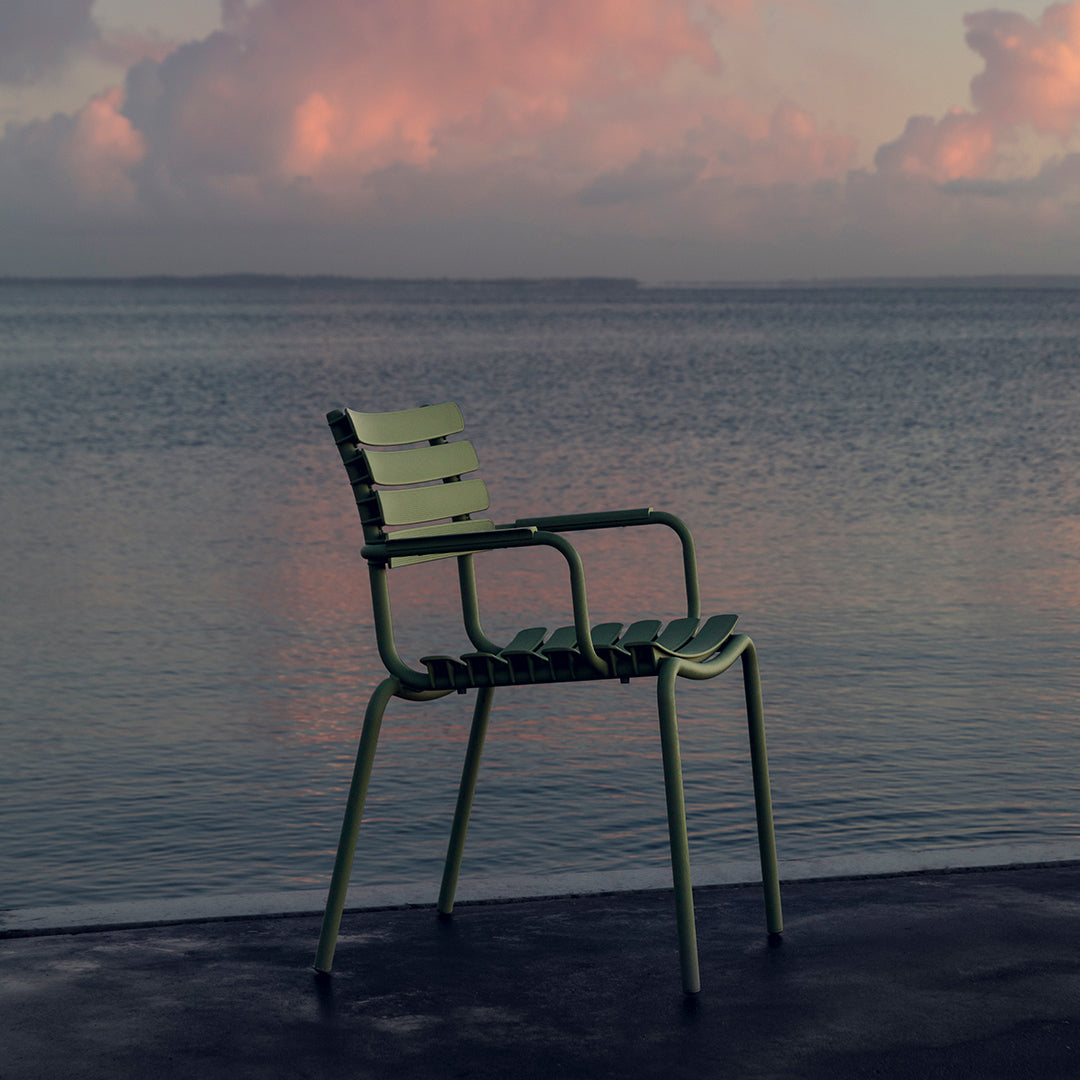 Green metal chair with slatted design, set against a serene water backdrop and colorful cloudy sky. 