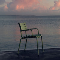 Green metal chair with slatted design, set against a serene water backdrop and colorful cloudy sky. 
