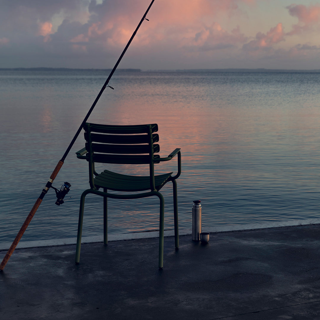 Green and black striped chair by calm water, reflecting soft pastel sunset hues.