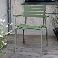 Green metal chair with horizontal slats, set against a rustic backdrop and surrounded by greenery.