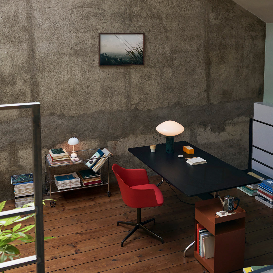 Modern workspace with red chair, black desk, wooden floor, and grey concrete wall, accented by a colorful art piece.