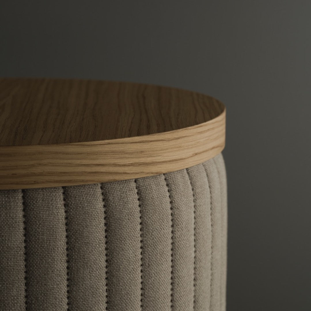 Beige upholstered stool with vertical stitching and a light oak top on a dark background.