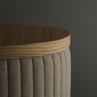 Beige upholstered stool with vertical stitching and a light oak top on a dark background.