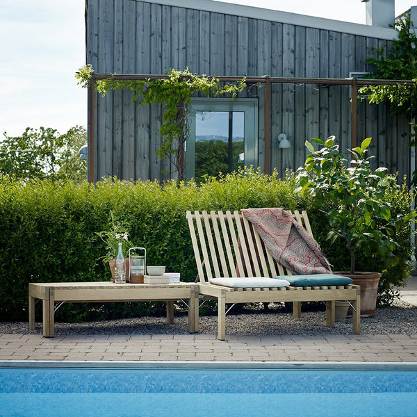 Light wooden lounge chair with slatted design; colorful patterned throw draped over the seat by a tranquil poolside.