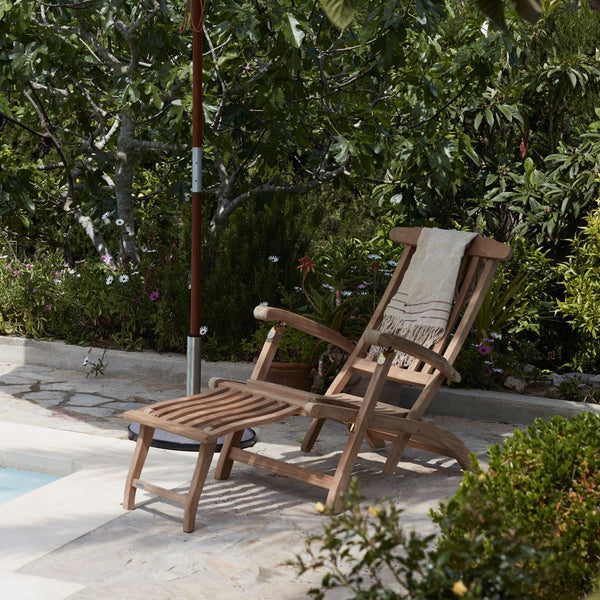 Natural wood reclining chair with a light-colored cushion, set against a lush green garden backdrop.