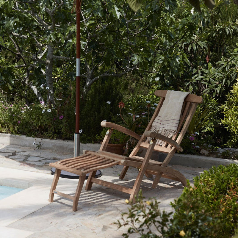 Natural wood reclining chair with a light-colored cushion, set against a lush green garden backdrop.