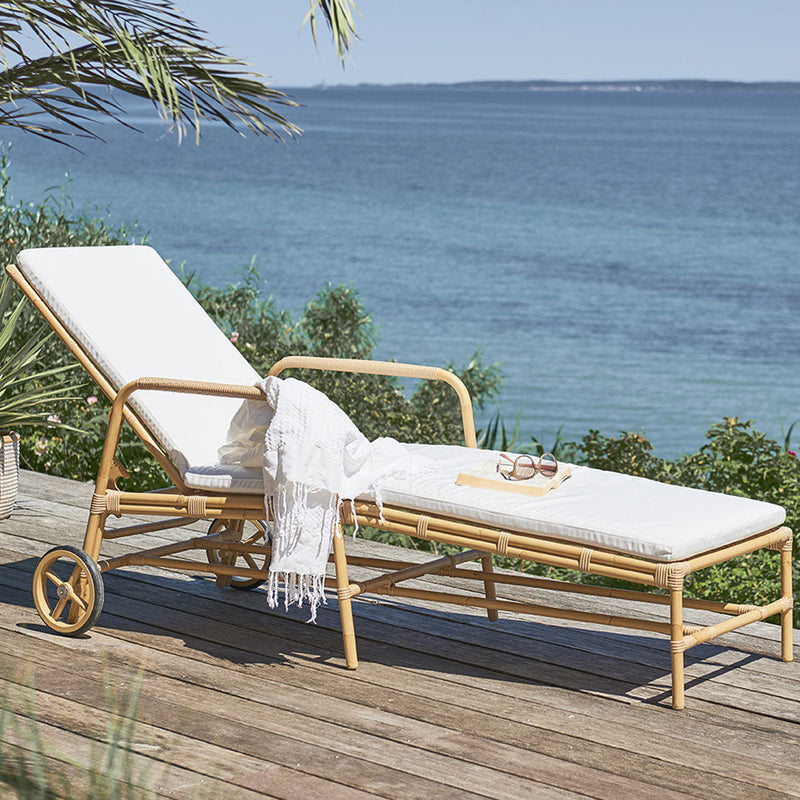 White cushioned lounge chair with natural rattan frame, set against a serene blue ocean backdrop. 