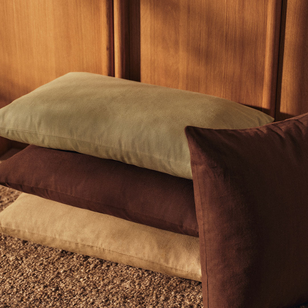 Stack of soft pillows in olive green, beige, and rich brown hues, against a wooden backdrop.