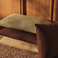 Stack of soft pillows in olive green, beige, and rich brown hues, against a wooden backdrop.