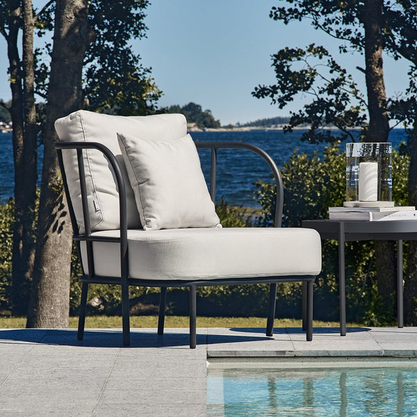Light beige cushion chair with a sleek black frame, set against a serene blue water backdrop and green foliage.