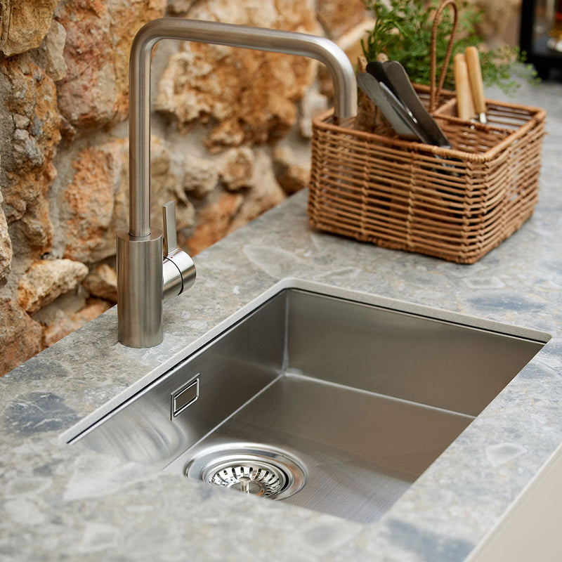 Stainless steel sink with a smooth, shiny finish, set against a textured stone countertop. Natural tones and earthy colors.