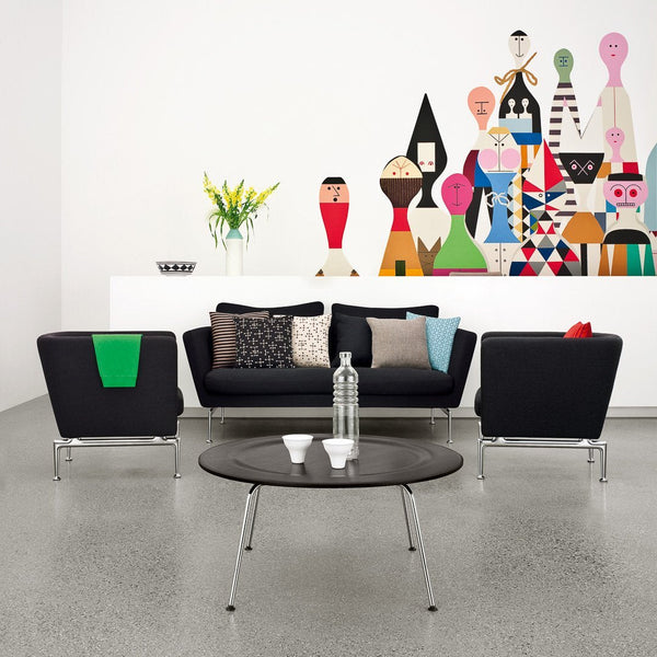 "Modern living room with black furniture, colorful patterned cushions, and a vibrant wall mural featuring abstract figures."