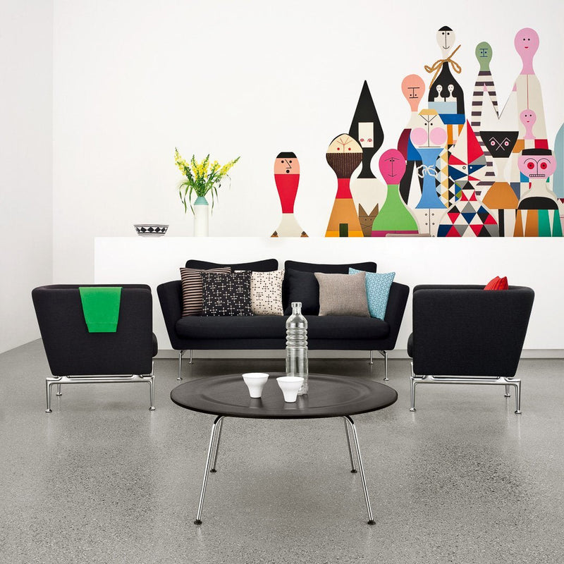 "Modern living room with black furniture, colorful patterned cushions, and a vibrant wall mural featuring abstract figures."