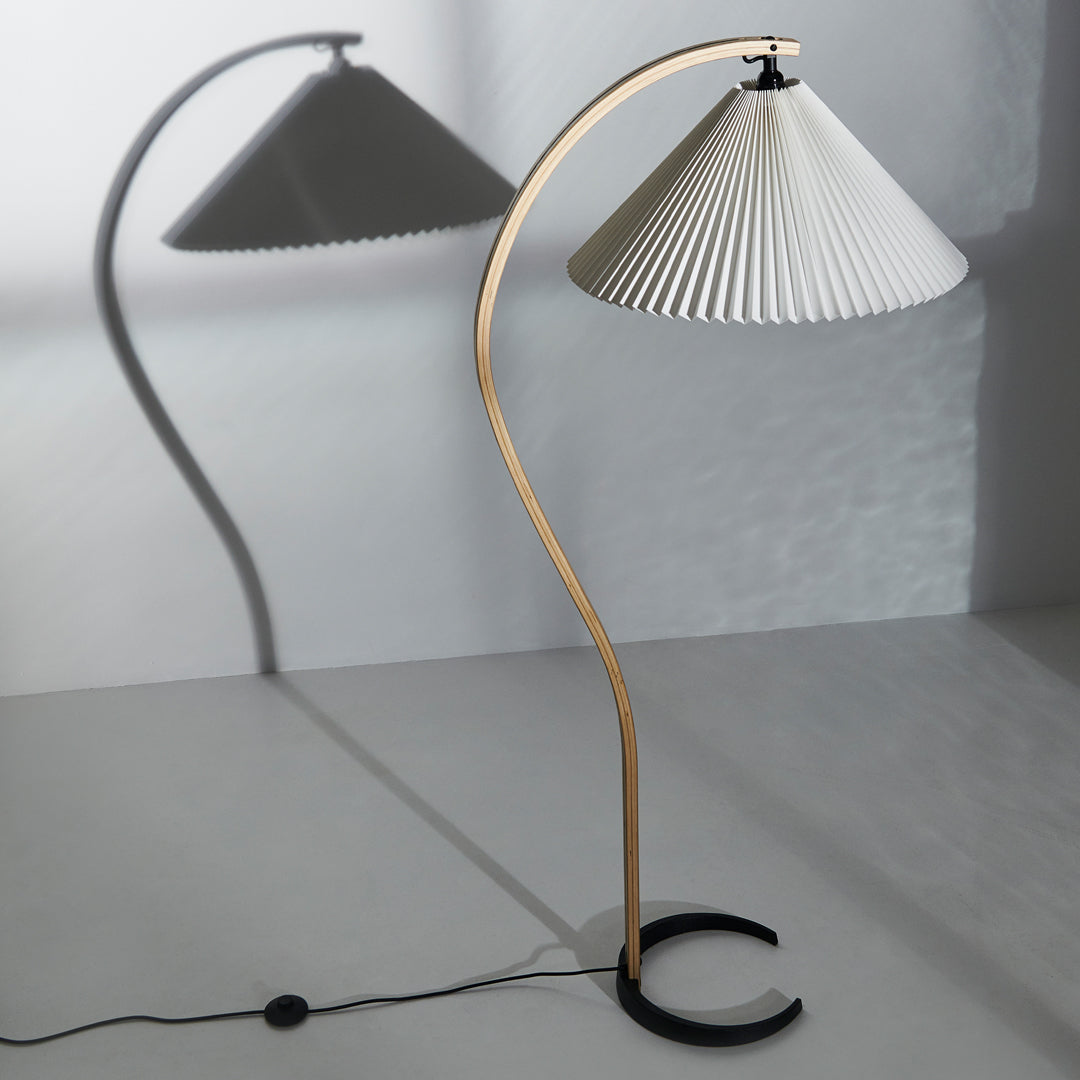 Elegant white pleated lamp shade with a light wood curved base and black circular stand, casting soft shadows.