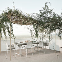 Elegant outdoor dining setup featuring white chairs and a natural woven canopy adorned with greenery and soft white blooms.