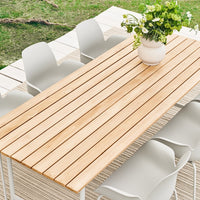 Light wood slatted dining table with six white minimalist chairs, featuring a fresh flower arrangement in a vase.