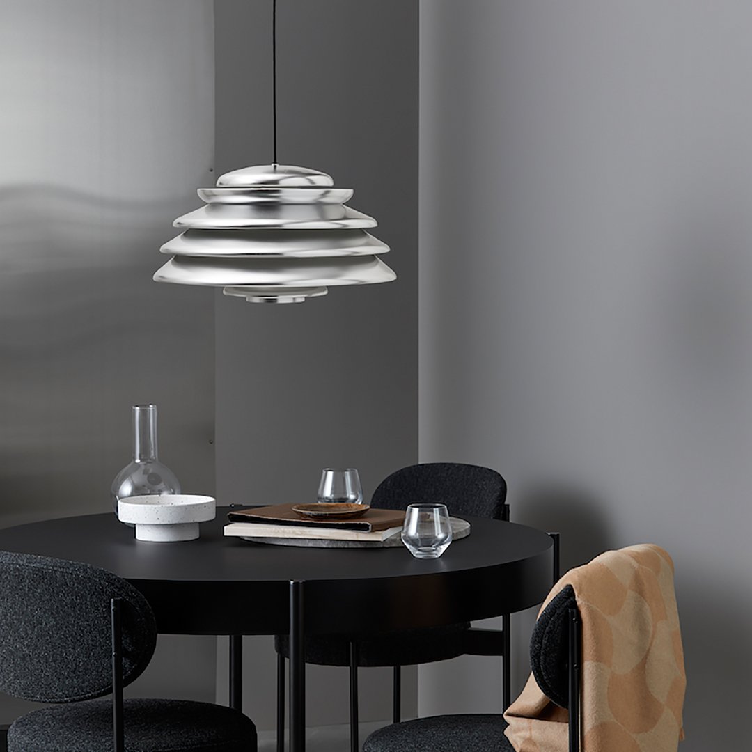 Modern silver pendant lamp with layered design, set against a neutral gray backdrop and a sleek black table.