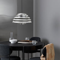 Modern silver pendant lamp with layered design, set against a neutral gray backdrop and a sleek black table.
