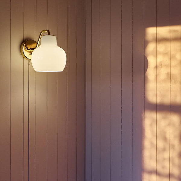 Elegant white glass wall lamp with a gold fixture casting soft light and a warm shadow on textured beige paneling.