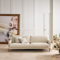 "Beige sofa with a smooth fabric, accented by a soft pillow, set against a neutral-toned background and floral artwork."