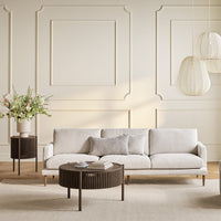 Beige modern sofa with gray accent pillow, paired with dark wood round table and light-colored decor in a neutral room.