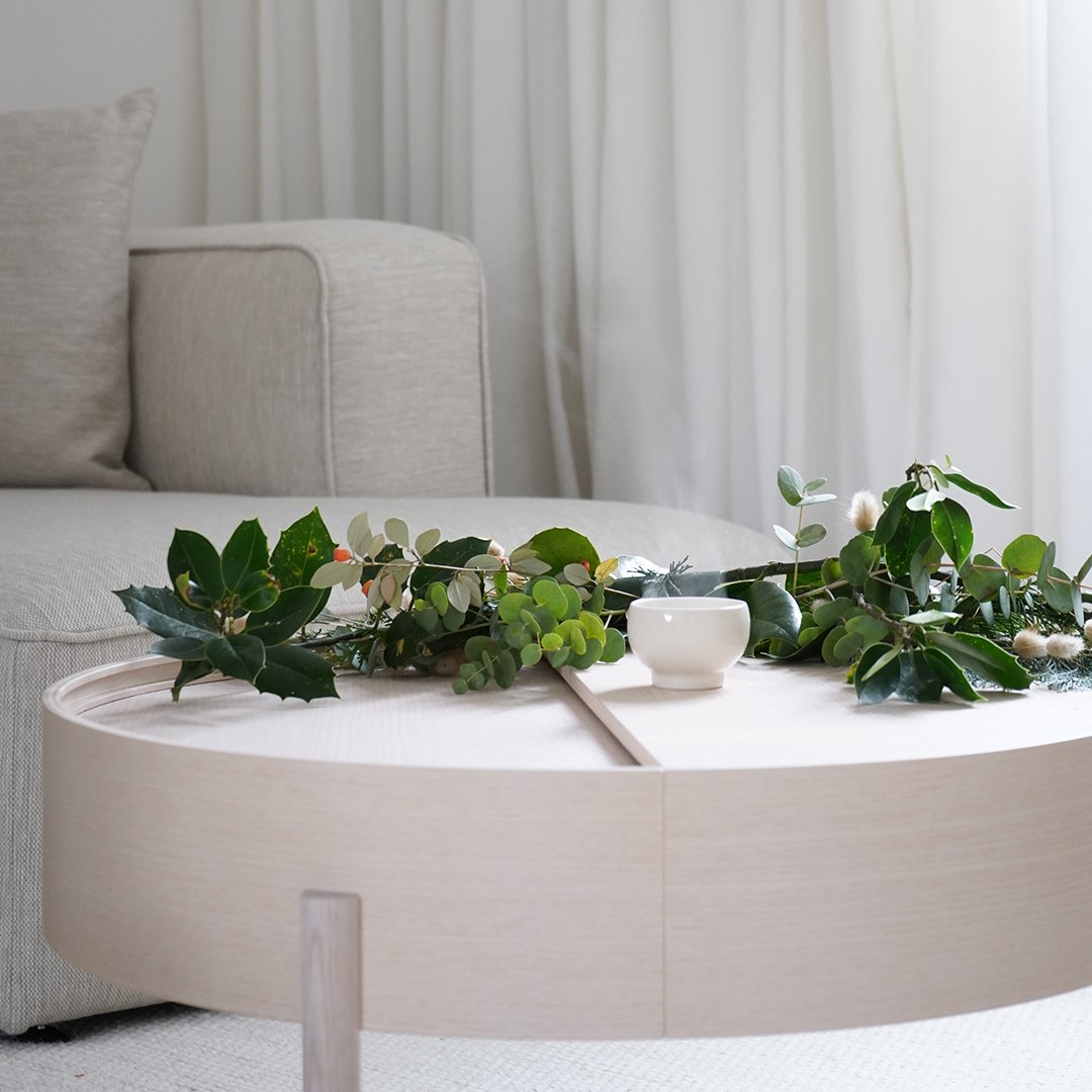 Neutral beige coffee table adorned with lush green foliage and a white ceramic bowl, set against a light, airy backdrop.