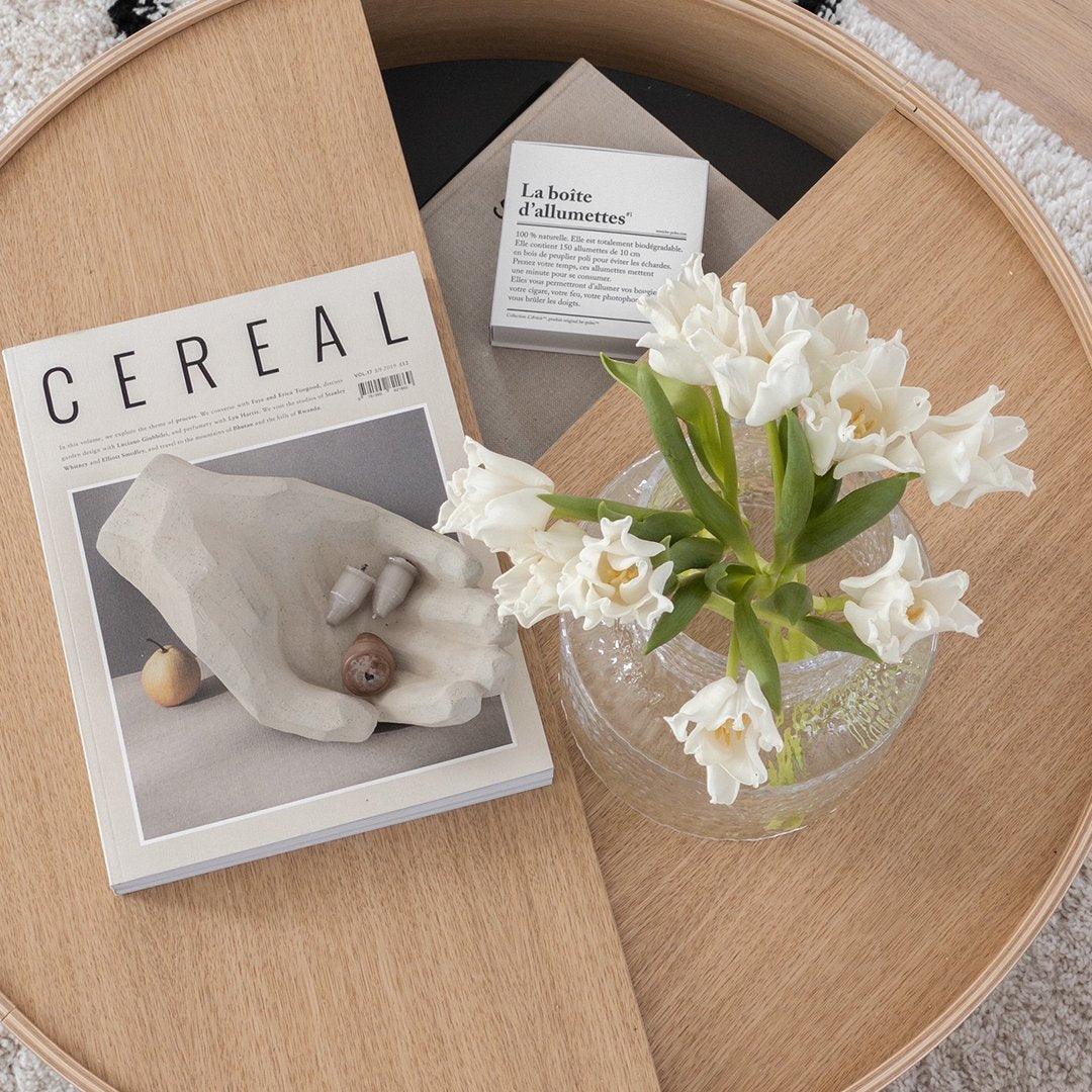 "White floral arrangement with a clear glass vase, on a wooden surface featuring a magazine with a minimalist design."