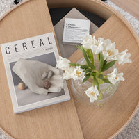 "White floral arrangement with a clear glass vase, on a wooden surface featuring a magazine with a minimalist design."