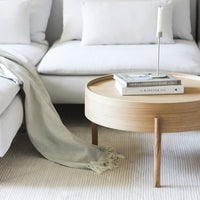 Light wood round coffee table with neutral beige throw and white sofa, featuring minimalist patterns and soft textures.