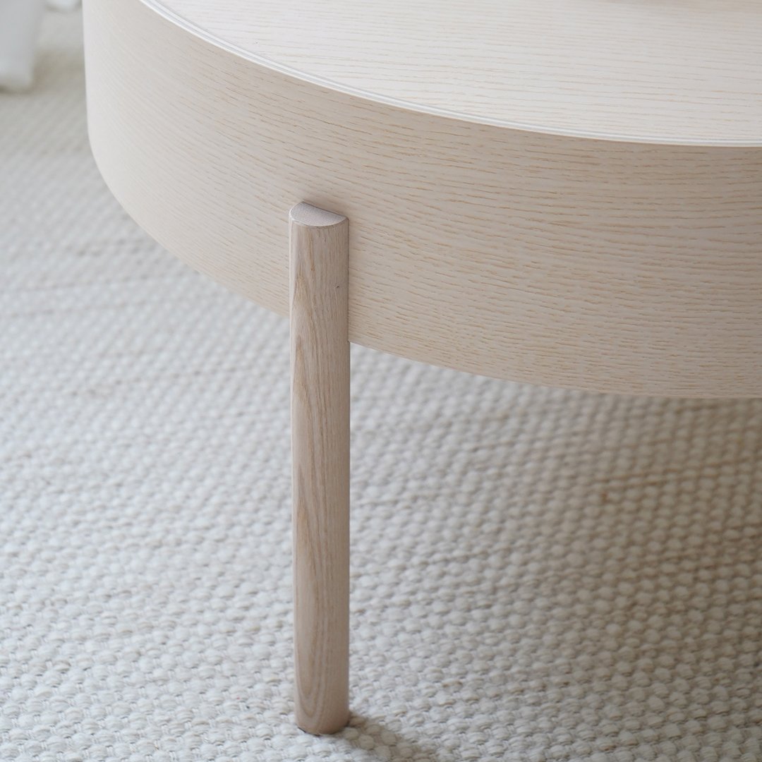 Light wood round coffee table with minimalistic design and smooth, pale finish, set against textured, neutral area rug.