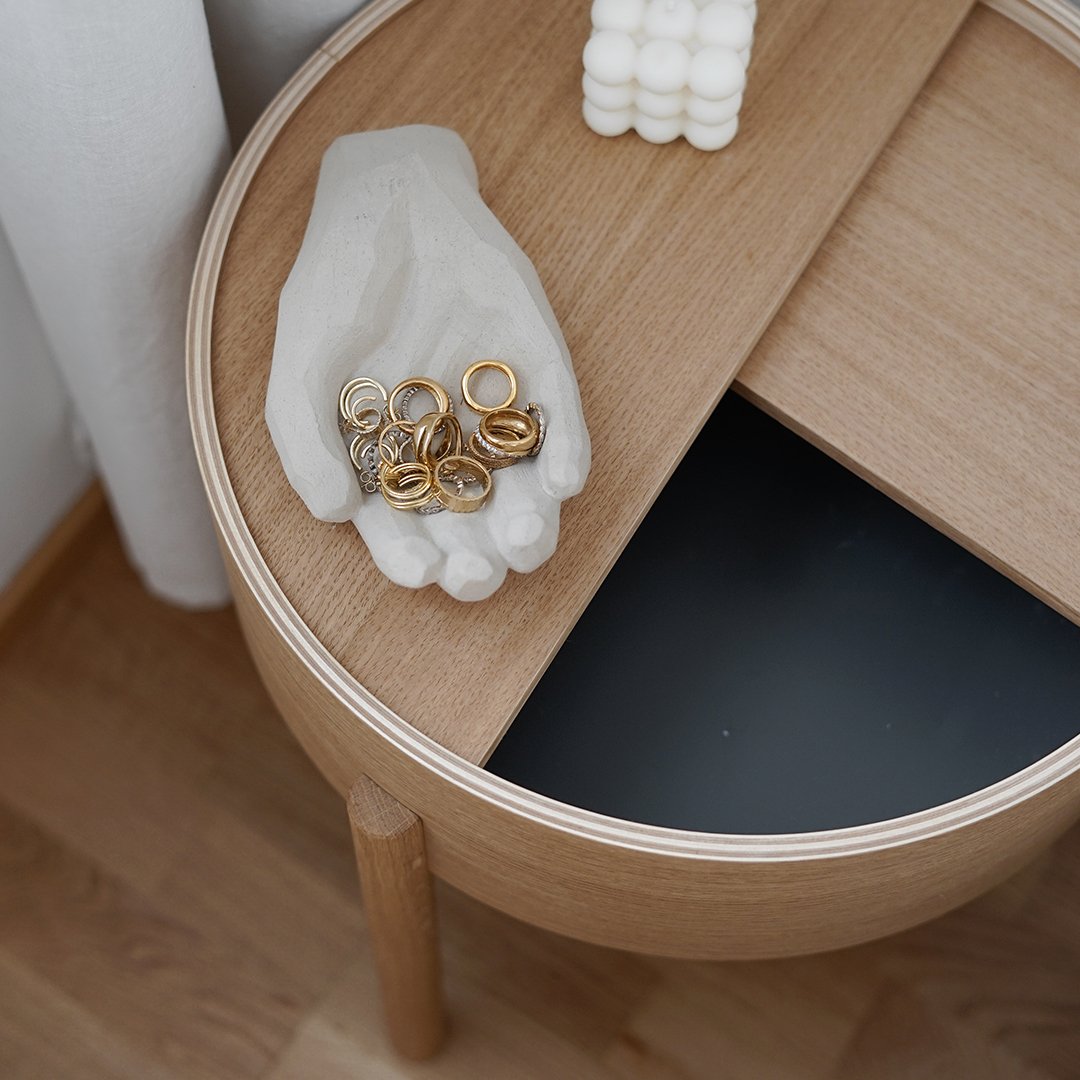 "Round wooden table with light wood finish, black inlay, and a textured beige hand holding gold rings."