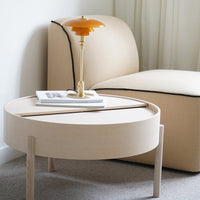 Round beige coffee table with natural wood legs, paired with a soft, light beige chair and a vibrant orange lamp.