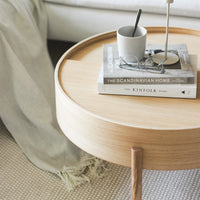 Light wood round side table with natural finish, featuring soft gray decor and patterned textile accents.
