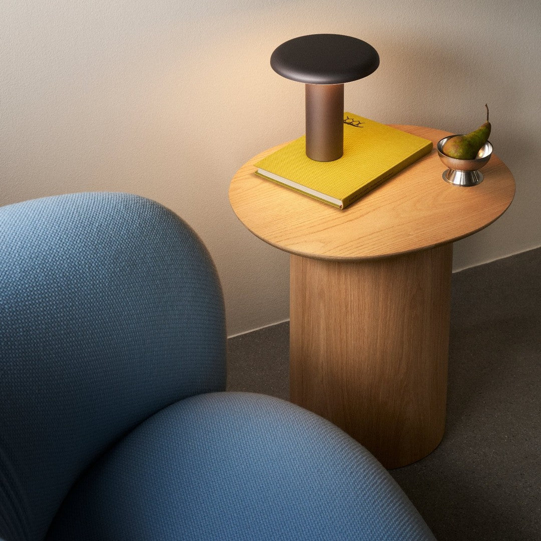 Cylindrical wooden side table with a smooth oak finish, complemented by a blue armchair and a yellow book.