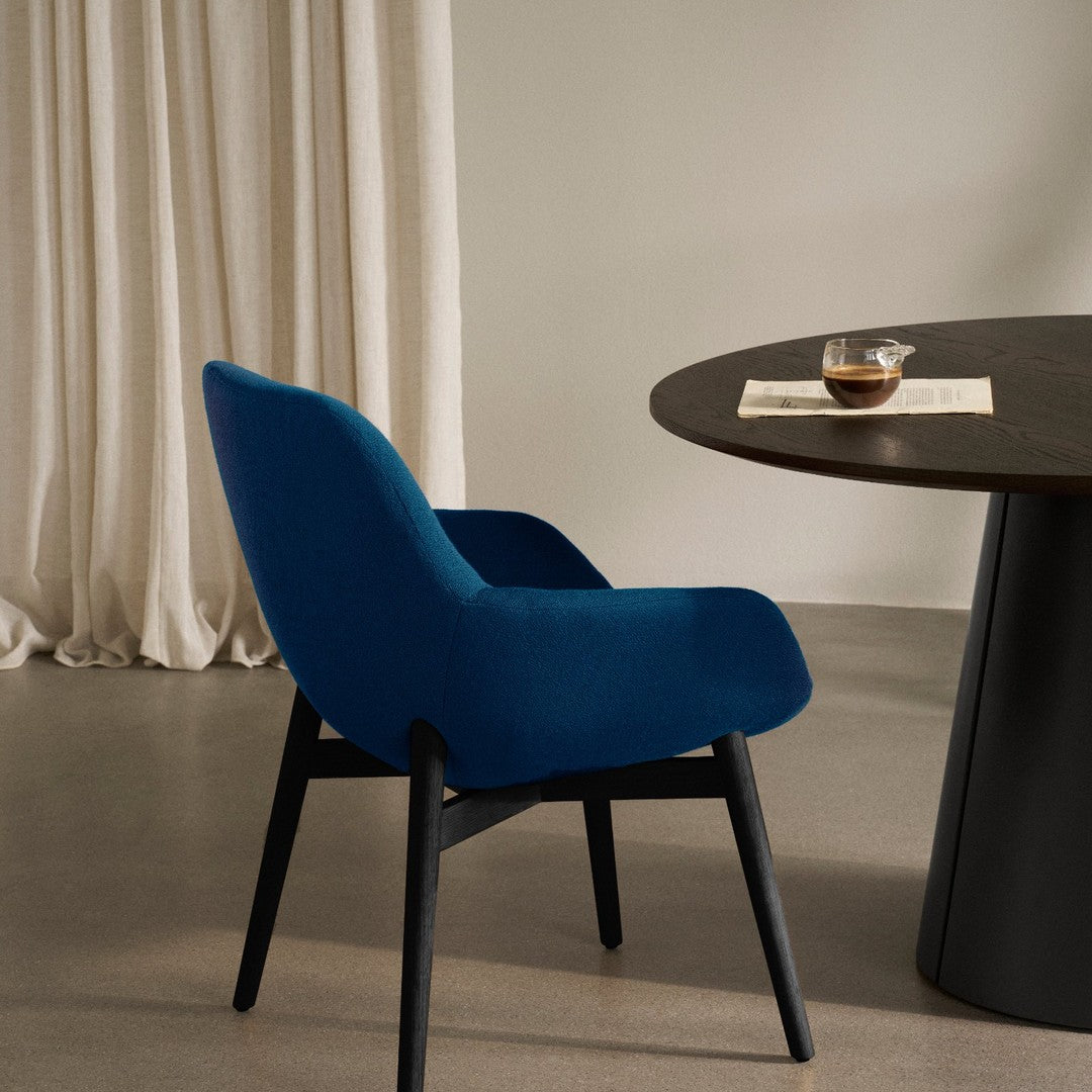 Blue velvet chair with sleek black legs next to a round dark table, complemented by soft cream curtains.