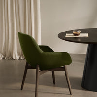 Green upholstered chair with wood legs beside a round dark table, set against soft beige curtains. Elegant minimalism.