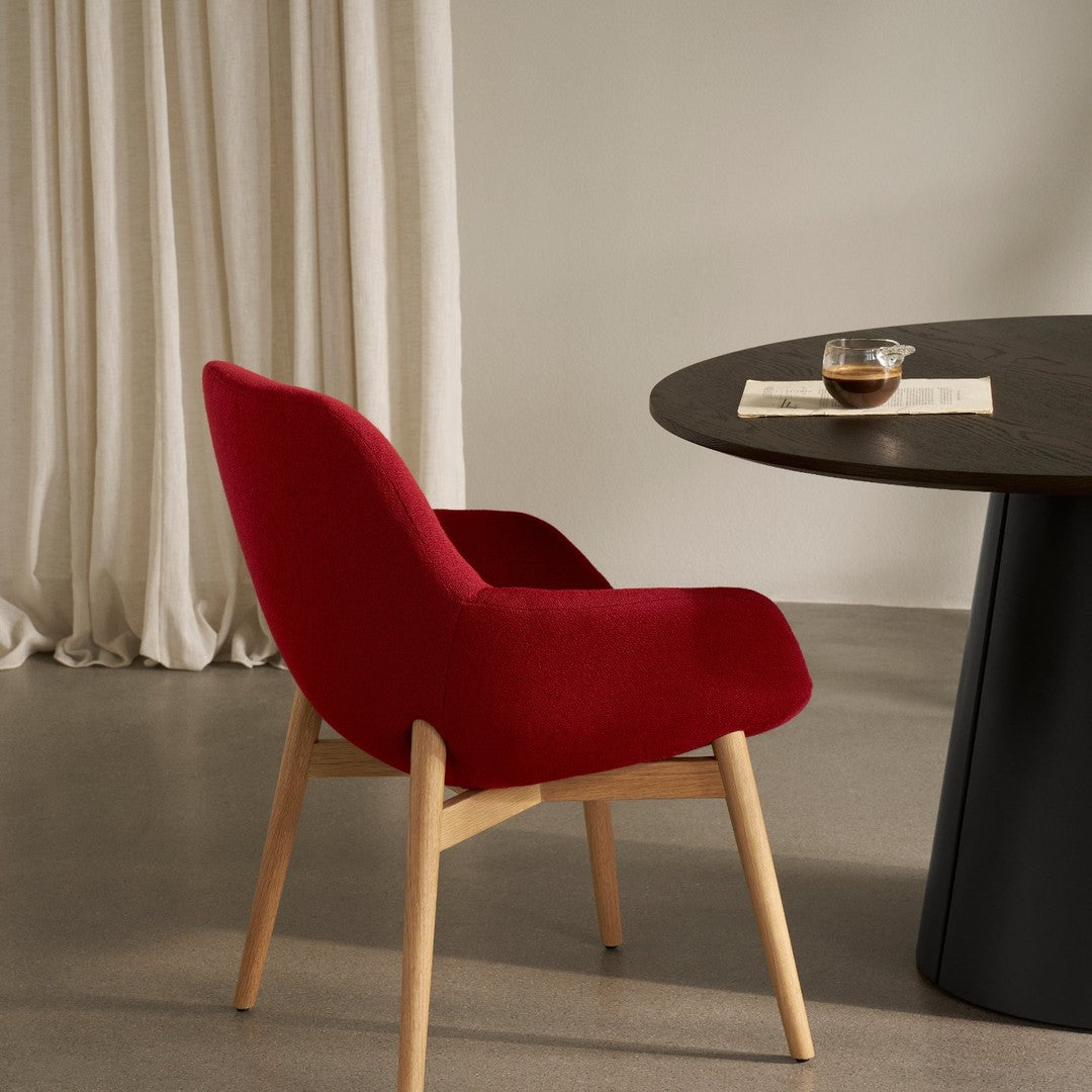 Red upholstered chair with curved back, paired with a round black table and soft white curtains in a minimalistic setting.