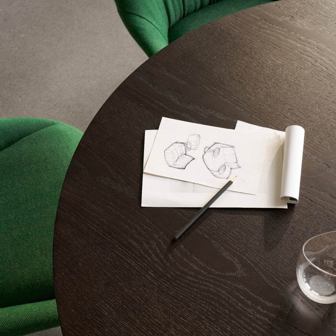 Green textured chairs surround a dark wooden table with sketches on paper, showcasing a minimalist and modern aesthetic.