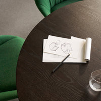 Green textured chairs surround a dark wooden table with sketches on paper, showcasing a minimalist and modern aesthetic.