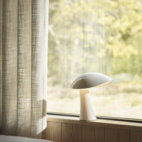 Minimalist white lamp with smooth curves, set against natural wood tones and soft beige curtains.