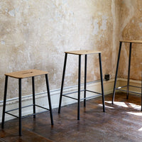 Wooden bar stools with natural wood tops and sleek black metal legs against a rustic, textured wall.