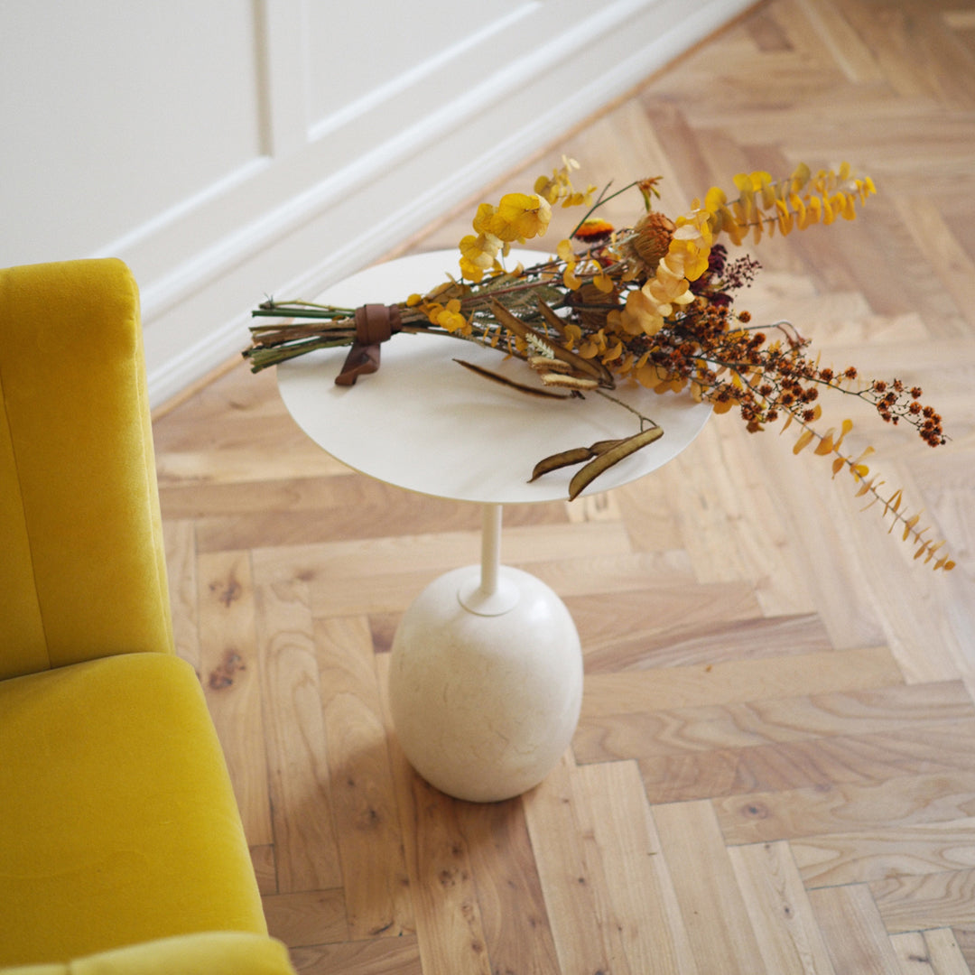 Minimalist white round tabletop on beige base, with vibrant yellow and orange floral arrangement, set against wooden flooring.