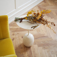 Minimalist white round tabletop on beige base, with vibrant yellow and orange floral arrangement, set against wooden flooring.