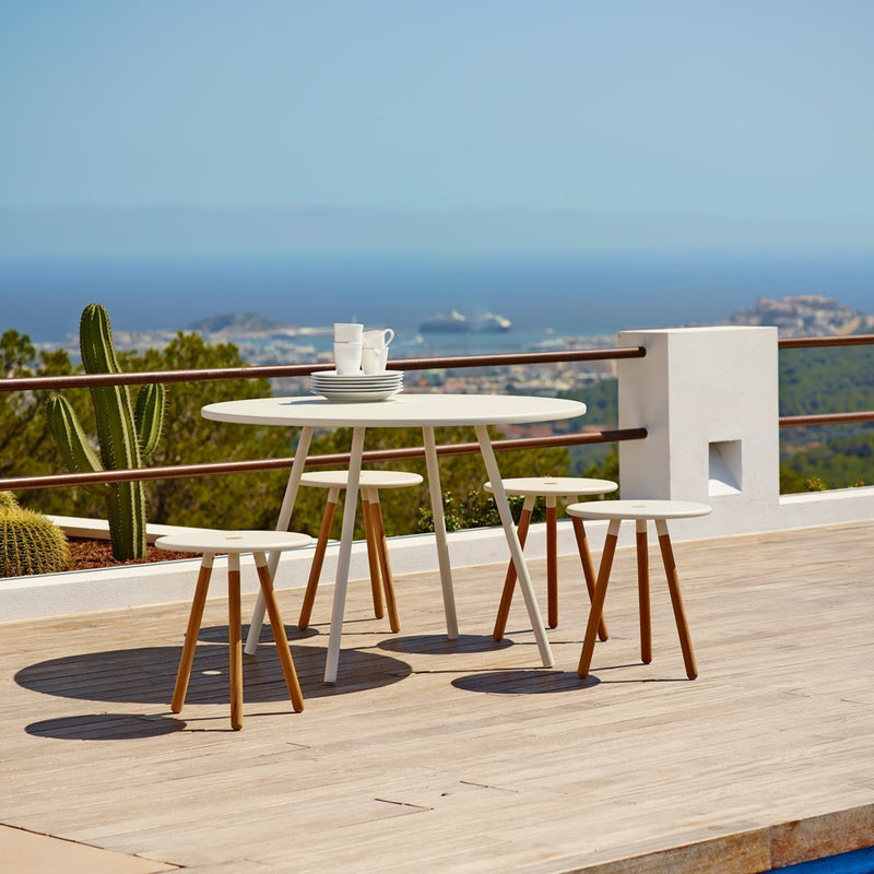 Stylish white outdoor table with natural wood legs, set against a serene blue ocean view and lush green plants.