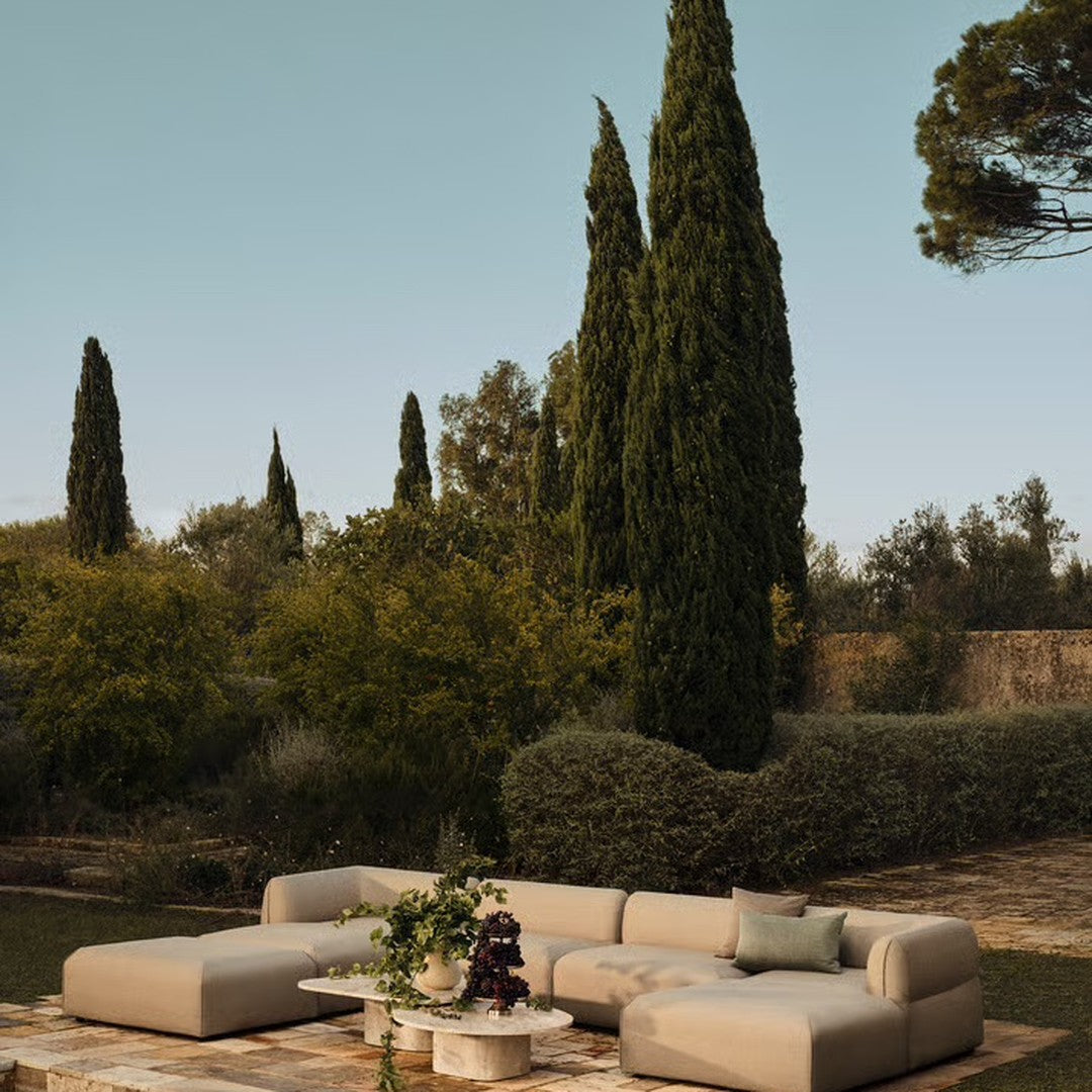 Beige sectional sofa with soft curves, set against lush greenery and tall cypress trees under a clear blue sky.