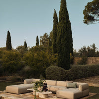 Beige sectional sofa with soft curves, set against lush greenery and tall cypress trees under a clear blue sky.