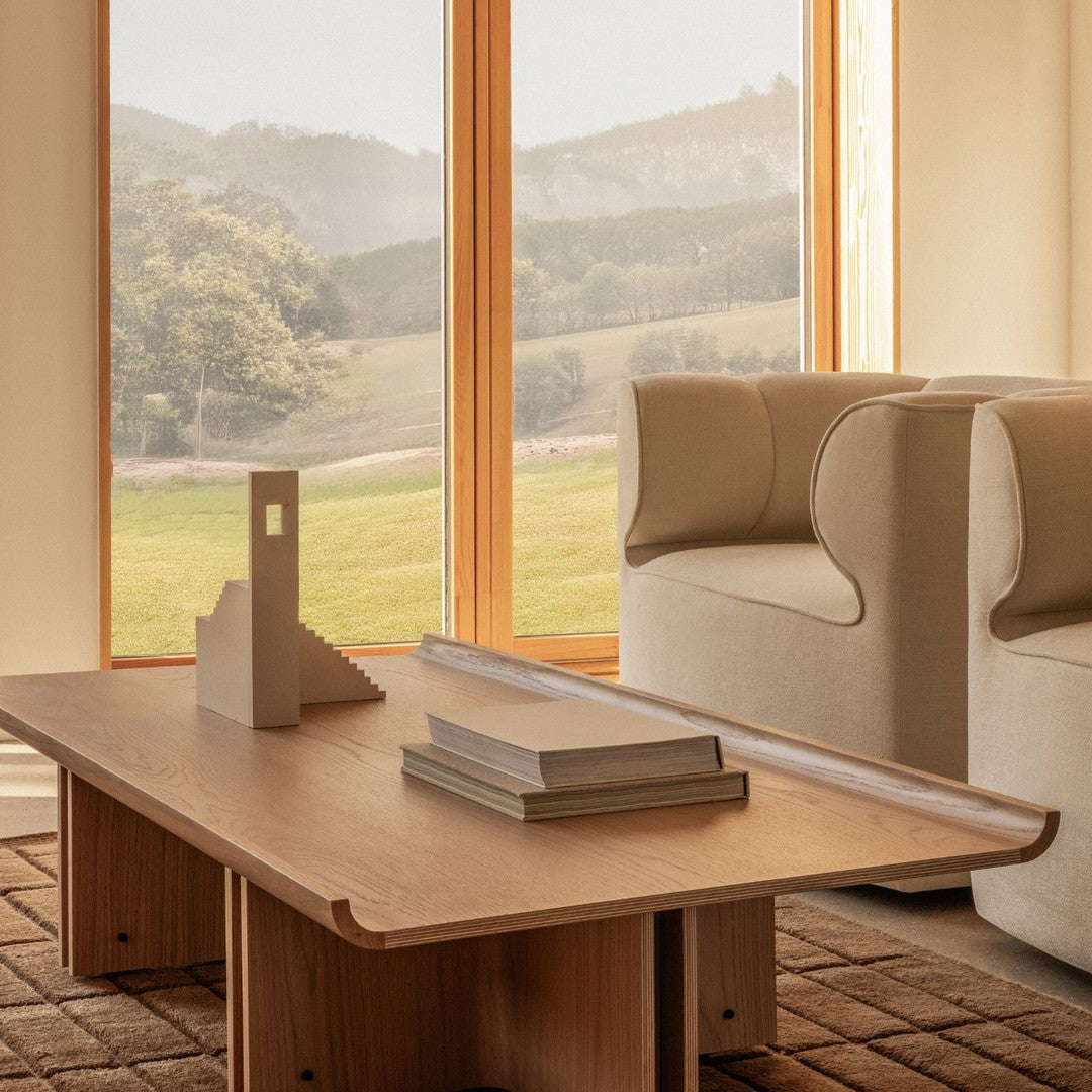 Light wood coffee table with smooth edges, set against a neutral-toned sofa and a textured brown rug. Natural light ambiance.