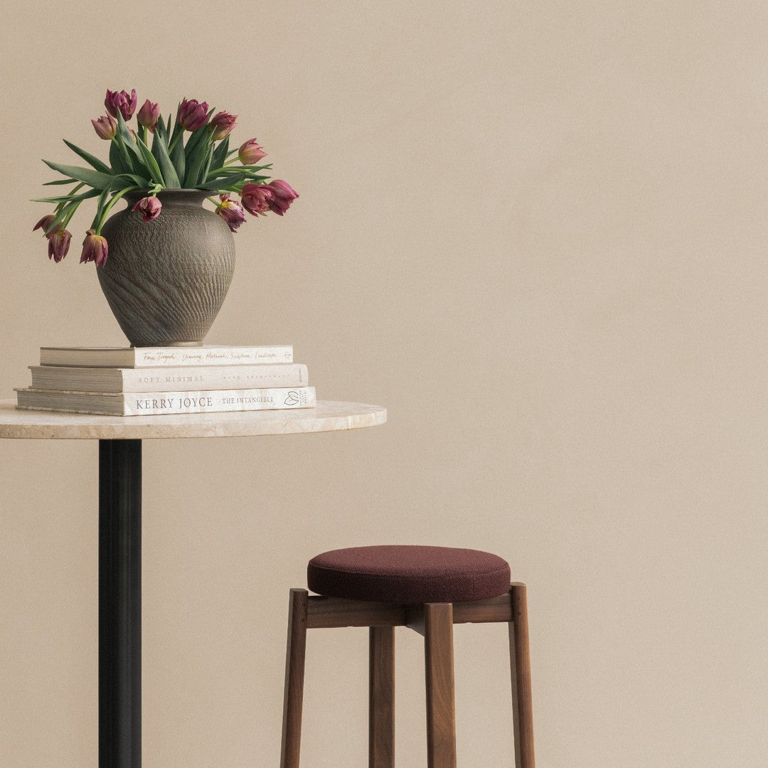 "Brown vase with green leaves and purple tulips, beige wall background, round cream table, and brown stool."