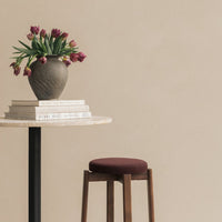 "Brown vase with green leaves and purple tulips, beige wall background, round cream table, and brown stool."