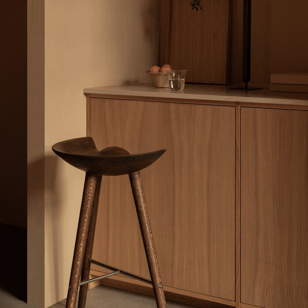 Wooden bar stool with a curved seat, set against a warm beige background and rich wood cabinetry.